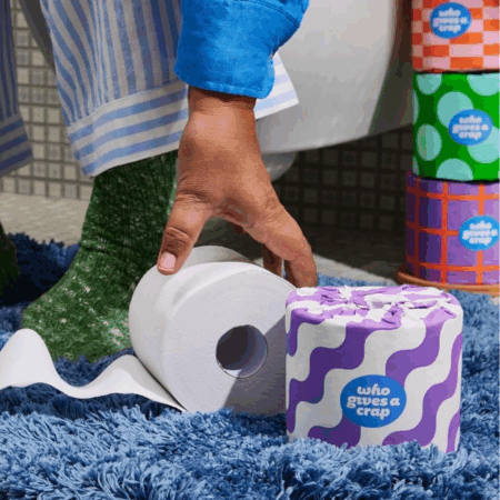 This image shows a hand reaching for a roll of toilet paper. The toilet paper is on a blue fuzzy rug. There are also several colorful rolls of toilet paper stacked behind the hand. The rolls of toilet paper are from the brand "Who Gives A Crap". The brand is known for its eco-friendly and sustainable toilet paper. The rolls are decorated with fun patterns like purple and white waves, orange and pink checkers, and green polka dots. This image is likely promoting sustainable bathroom products and eco-conscious living.