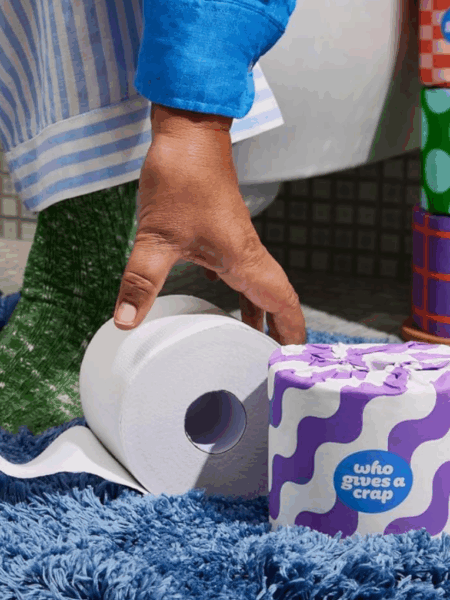 This image shows a hand reaching for a roll of toilet paper. The toilet paper is on a blue fuzzy rug. There are also several colorful rolls of toilet paper stacked behind the hand. The rolls of toilet paper are from the brand "Who Gives A Crap". The brand is known for its eco-friendly and sustainable toilet paper. The rolls are decorated with fun patterns like purple and white waves, orange and pink checkers, and green polka dots. This image is likely promoting sustainable bathroom products and eco-conscious living.
