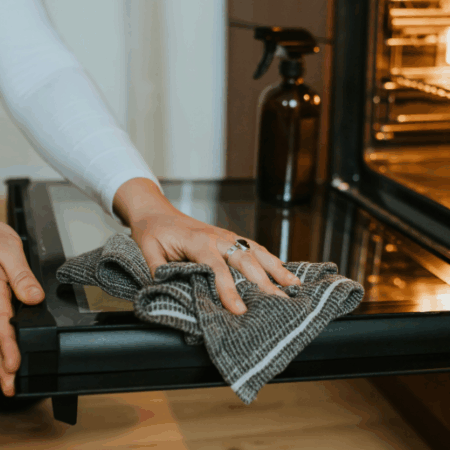 Person wiping down an open oven door with a cloth while using natural cleaning supplies.