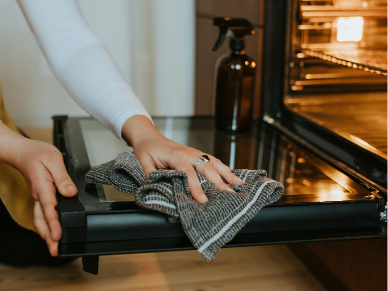 Person wiping down an open oven door with a cloth while using natural cleaning supplies.