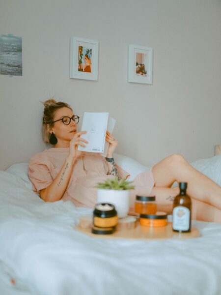 Woman relaxing on a bed while reading a book, wearing glasses and neutral loungewear, with skincare products and a small plant on a tray in the foreground in a calm, minimalist bedroom.