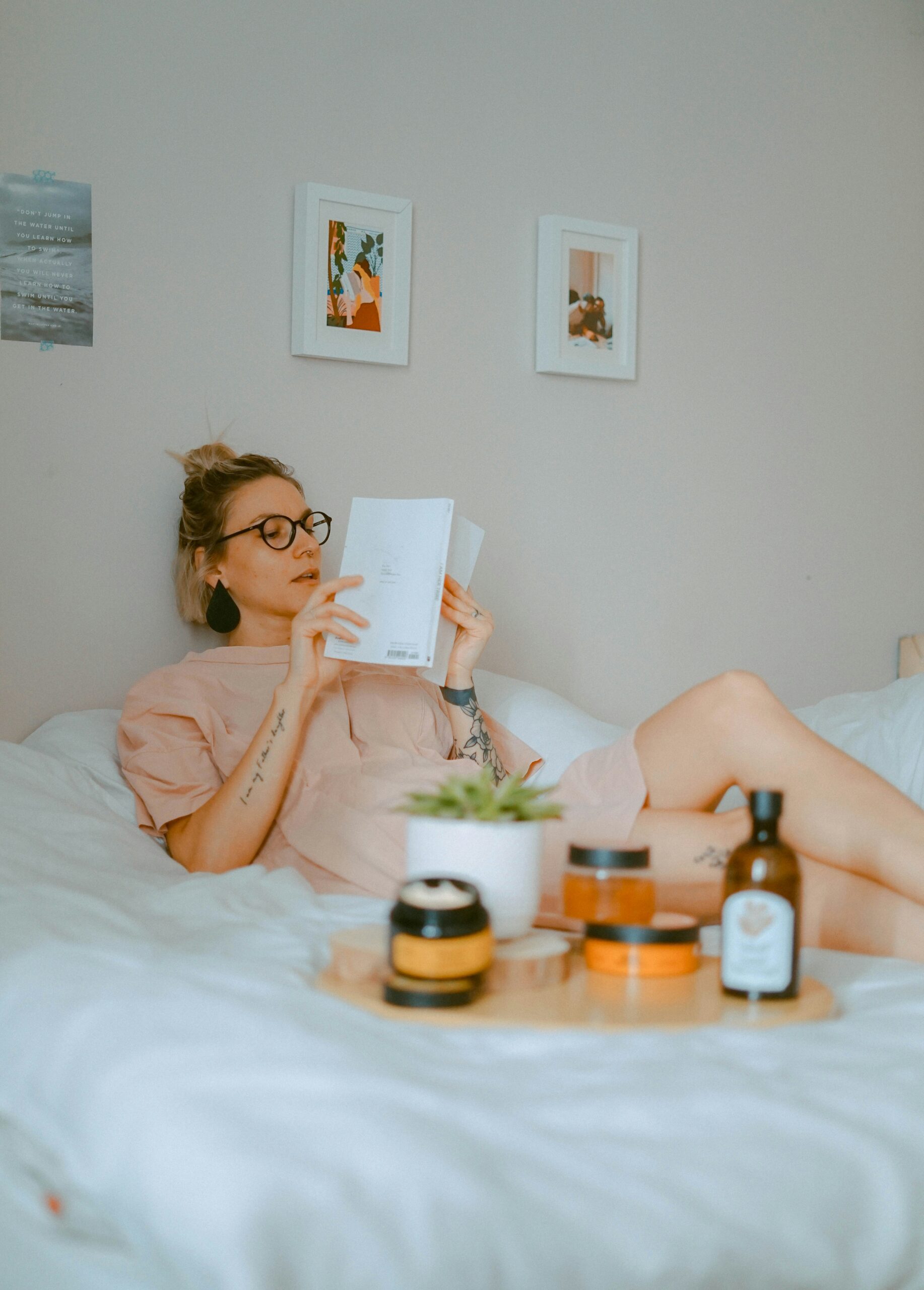 Woman relaxing on a bed while reading a book, wearing glasses and neutral loungewear, with skincare products and a small plant on a tray in the foreground in a calm, minimalist bedroom.