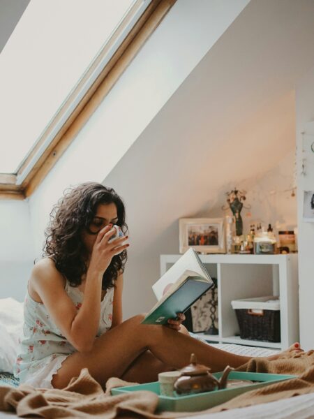 Woman sitting indoors in natural daylight, holding a book and sipping a drink, pausing quietly in a calm, lived-in home setting.