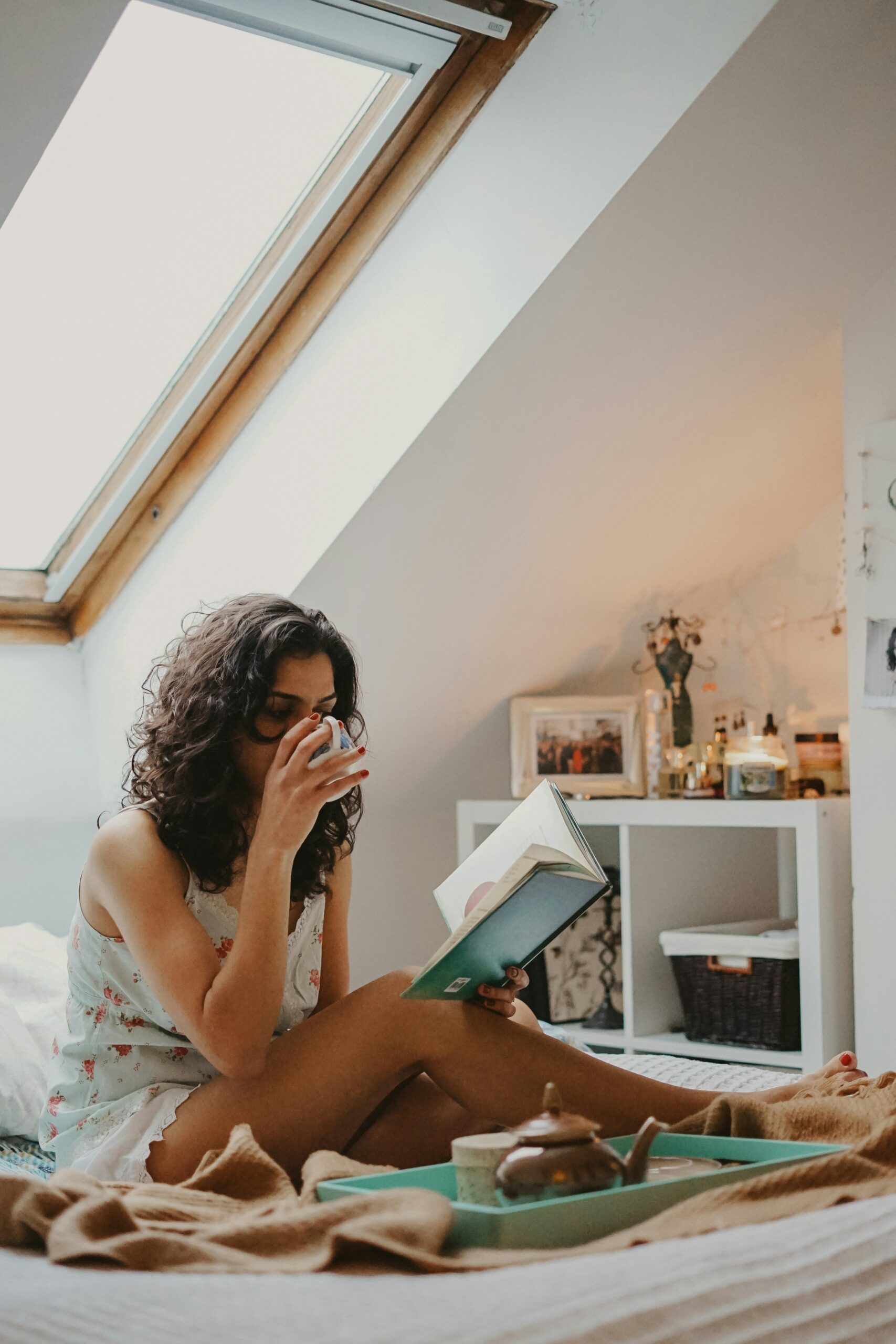 Woman sitting indoors in natural daylight, holding a book and sipping a drink, pausing quietly in a calm, lived-in home setting.