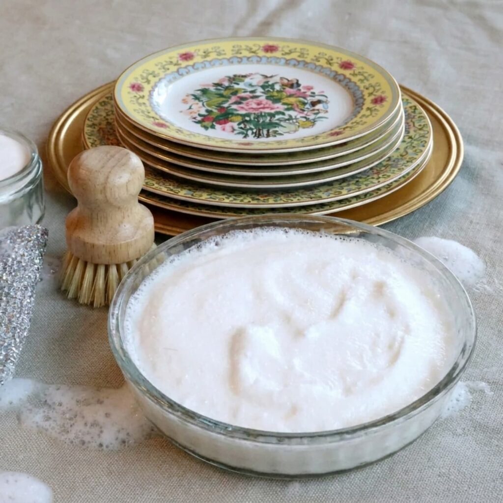 Flower dishes in the background with homemade dish soap in a glass bowl and a wooden dish brush.
