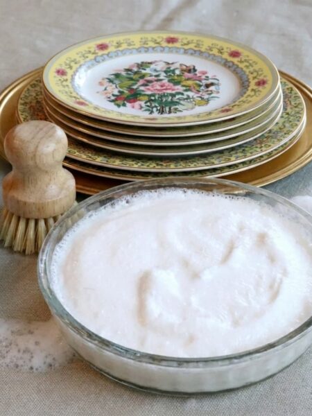 Flower dishes in the background with homemade dish soap in a glass bowl and a wooden dish brush.
