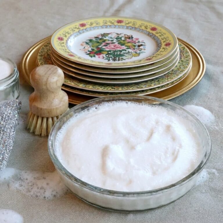 Flower dishes in the background with homemade dish soap in a glass bowl and a wooden dish brush.