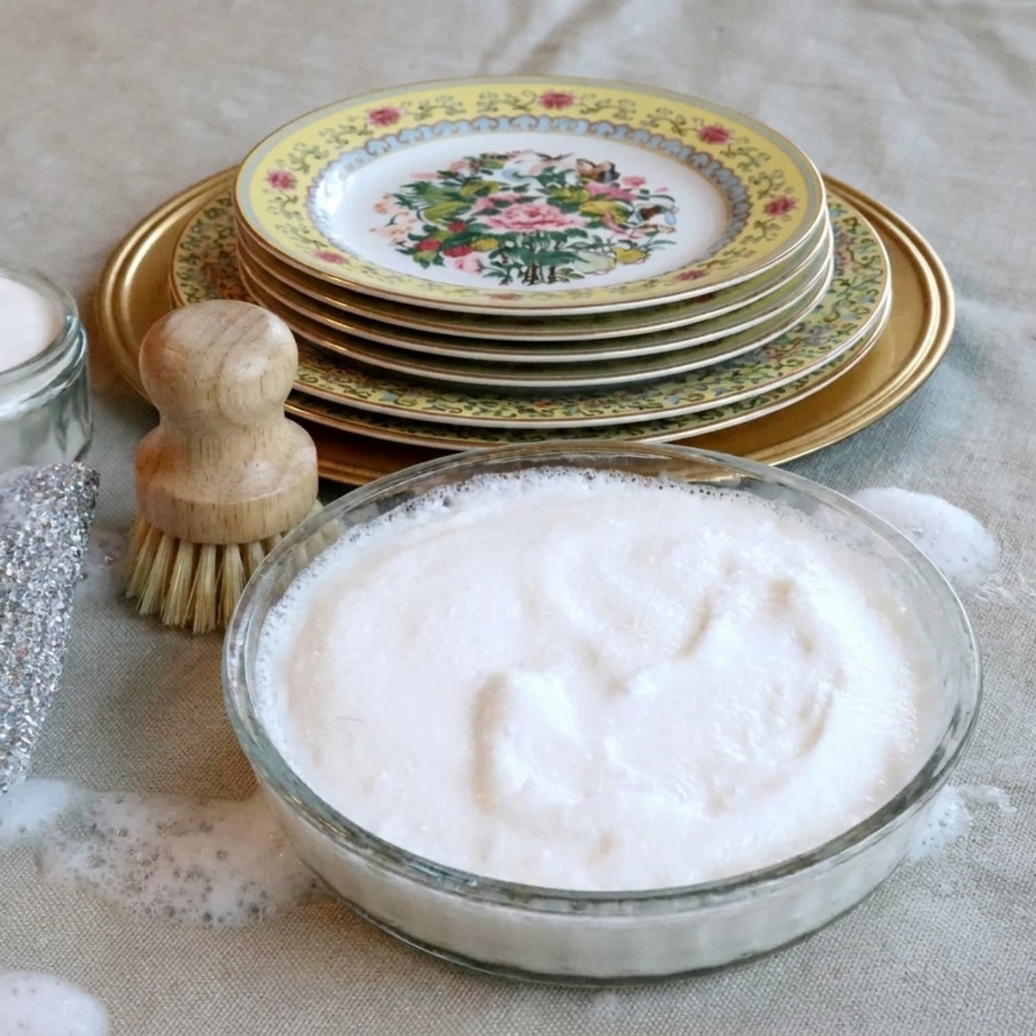 Flower dishes in the background with homemade dish soap in a glass bowl and a wooden dish brush.