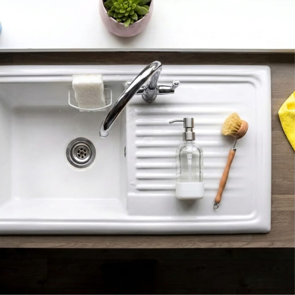 Overhead view of a clean white kitchen sink and drain with a glass soap dispenser and wooden dish brush, the kind of drain a homemade drain cleaner keeps clear.