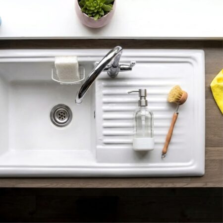 Overhead view of a clean white kitchen sink and drain with a glass soap dispenser and wooden dish brush, the kind of drain a homemade drain cleaner keeps clear.