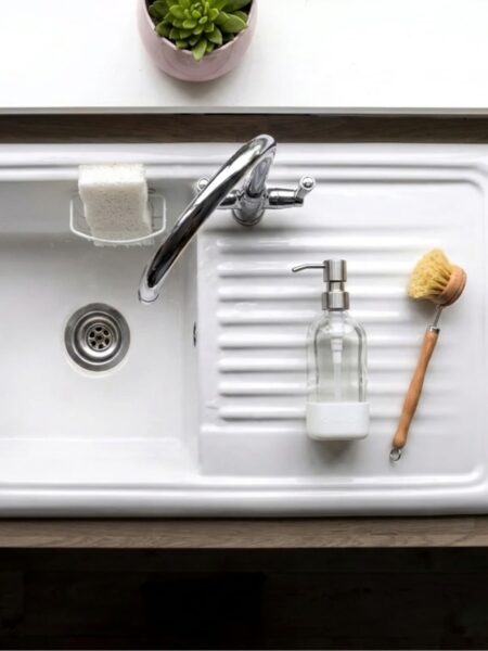 Overhead view of a clean white kitchen sink and drain with a glass soap dispenser and wooden dish brush, the kind of drain a homemade drain cleaner keeps clear.