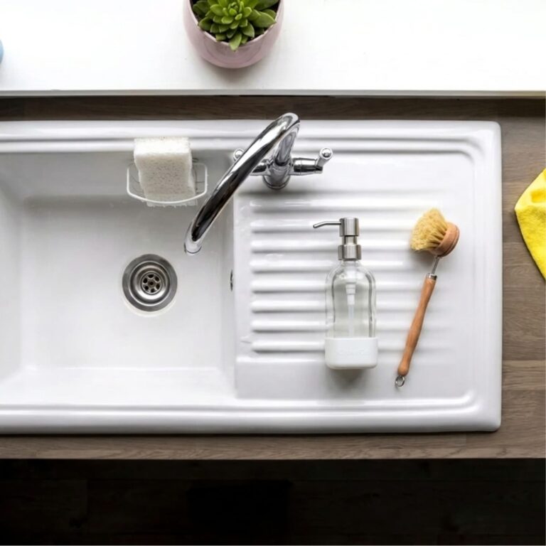 Overhead view of a clean white kitchen sink and drain with a glass soap dispenser and wooden dish brush, the kind of drain a homemade drain cleaner keeps clear.