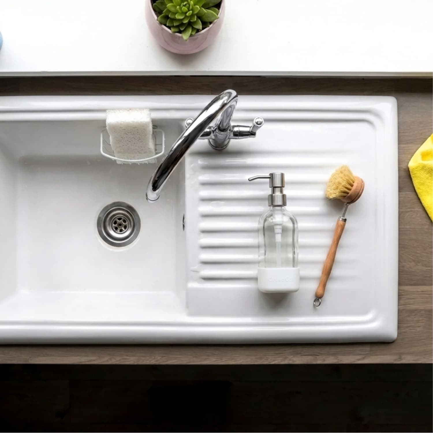 Overhead view of a clean white kitchen sink and drain with a glass soap dispenser and wooden dish brush, the kind of drain a homemade drain cleaner keeps clear.