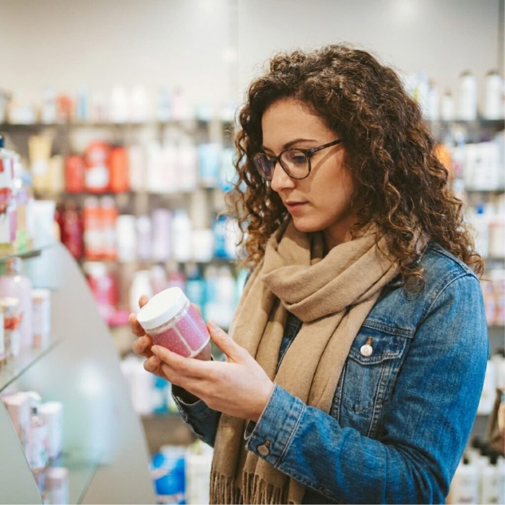 Woman with curly hair and glasses reading the ingredient label on a skincare product jar in a store aisle.