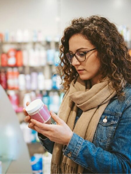 Woman with curly hair and glasses reading the ingredient label on a skincare product jar in a store aisle.