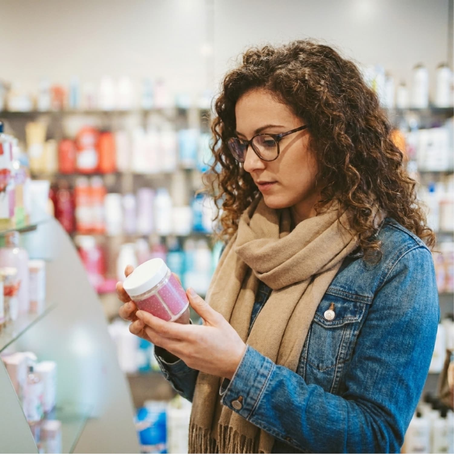 Woman with curly hair and glasses reading the ingredient label on a skincare product jar in a store aisle.