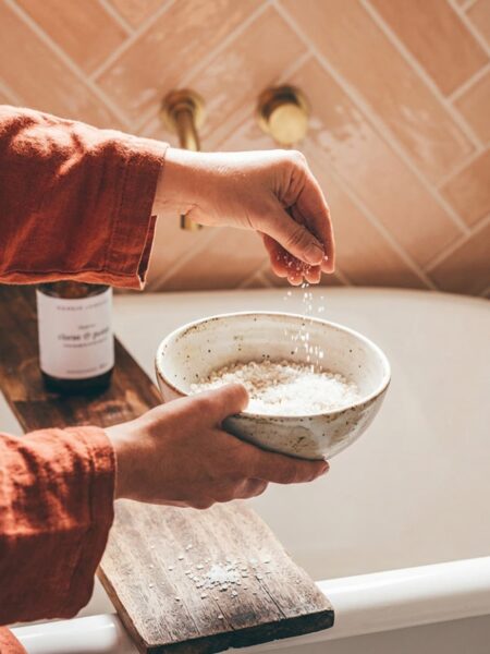 Hands in linen sleeves sprinkling bath salts from a speckled ceramic bowl over a bathtub, a simple self-care ritual