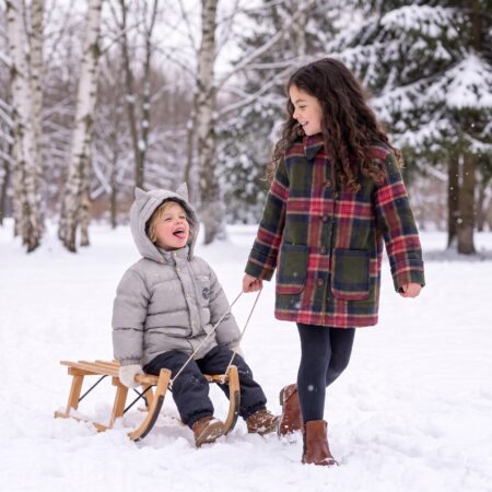 Best kids winter coats, a girl in a plaid wool coat pulling her little brother in a cat-ear puffer on a sled through the snow