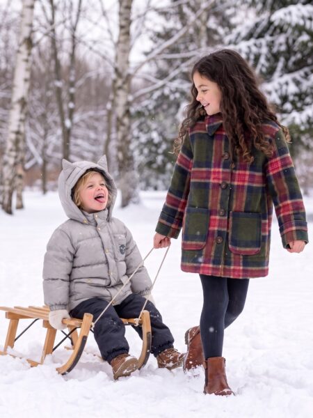 Best kids winter coats, a girl in a plaid wool coat pulling her little brother in a cat-ear puffer on a sled through the snow