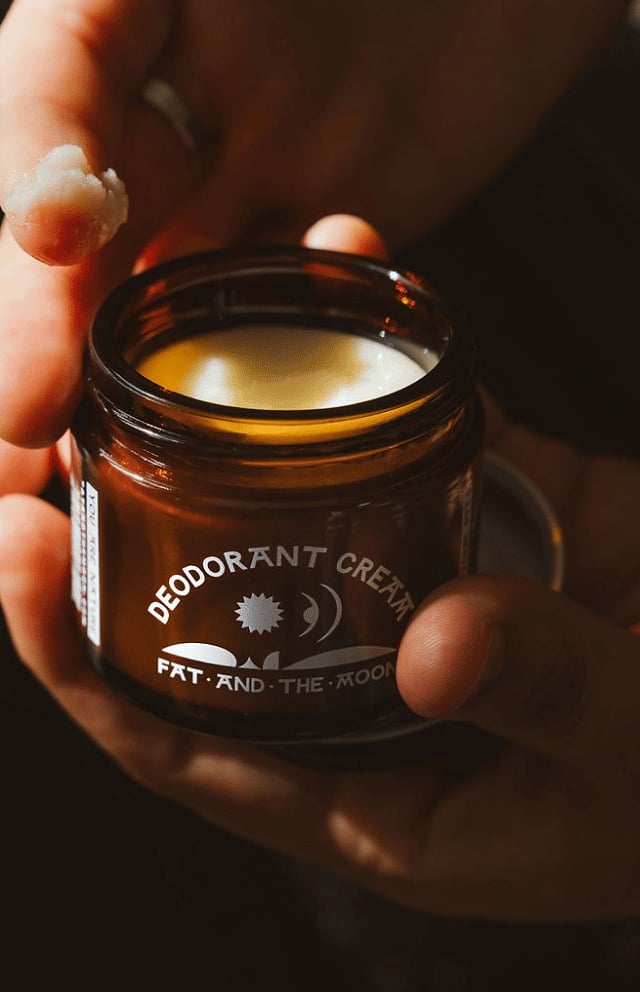 Hands holding an open amber glass jar of Fat and the Moon deodorant cream